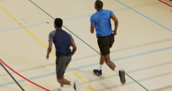Diverse male athletes jogging then dribbling ball toward hoop and rebounding on gym court