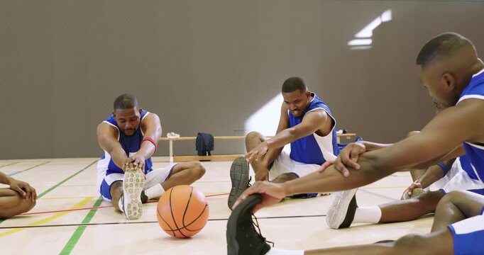 Diverse male players stretching hamstrings on court around basketball with towel before practice