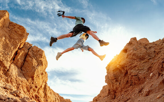 Adventurous hikers jumping over a rocky canyon gap in a desert landscape with blue sky and copy space.