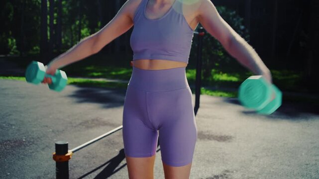 Fit anonymous young woman performing lateral raises with dumbbells during an outdoor workout session