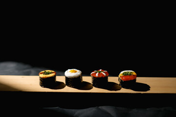 Row of four assorted creative Japanese fusion sushi rolls with various colorful toppings served on a long wooden board against a dark dramatic background. © Vlafy Studio