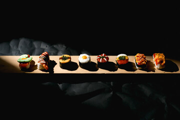 Long horizontal row of eight assorted creative Japanese fusion sushi pieces on a wooden board. Diverse colorful toppings featured against a dark dramatic crumpled paper background. © Vlafy Studio
