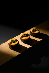 Three pieces of classic maki sushi roll topped with yellow mentai sauce and herbs. Arranged on a wooden board with a dramatic spotlight against a pitch black background. © Vlafy Studio