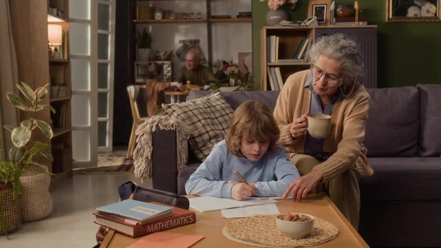 Long shot of senior Caucasian grandmother holding mug and helping child with mathematics homework at home. Footage is ideal for promoting homeschooling, family support, and senior lifestyle services