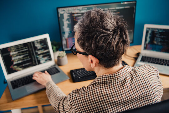 Programmer working on multiple laptops and monitors, writing code and debugging software at a home office desk