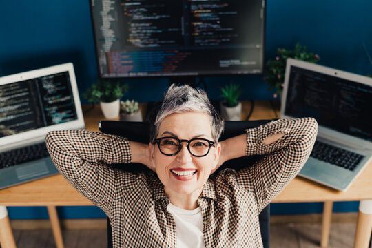 Smiling female programmer relaxing in a home office with laptops and code on screens, confident senior developer working remotely and enjoying productivity in a modern tech workspace