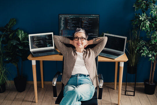 Relaxed woman programmer coding at home office desk with multiple laptops and monitor, smiling confidently while enjoying remote work, productivity and tech creativity
