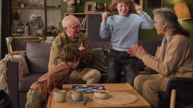 Medium long shot of Caucasian grandparents and grandchildren playing board game in living room, celebrating victory with high fives. Perfect for family bonding and intergenerational lifestyle