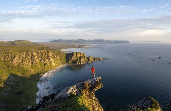 Evening mood, mountaineer standing on a rocky outcrop on the summit of M&aring;tind, view of coastal landscape, H&oslash;yvika beach and sea, M&aring;tinden, And&oslash;ya island, Vester&aring;len, Nordland Norway