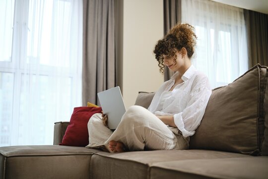 A young woman with curly hair sits comfortably on a couch, focused on her tablet in a bright, cozy living room. Soft curtains flutter with light, creating a serene atmosphere for reading
