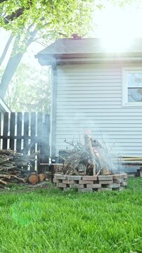 A wood pile sits beside a house with smoke coming from a fire. The sun shines brightly as green grass grows nearby. The fence adds a rustic look to the scene.
