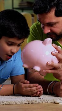 Indian asian young father son taking out coins from piggy bank with happiness while lying on carpet together, learning saving habits and enjoying joyful family bonding time in modern living room