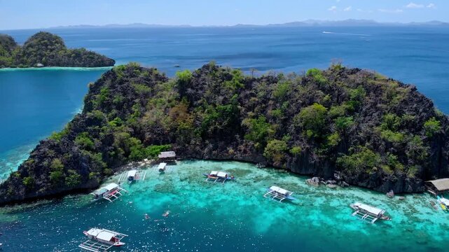 Overhead reveal of rugged karst island surrounded by crystal clear turquoise shallows with traditional outrigger boats anchored along a sheltered cove. Coron, Palawan, Philippines