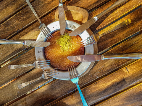 Turkish kunefe dessert with pistachio powder topping in metal plate on wooden table. Traditional sweet pastry food surrounded by fork and knife cutlery, top view, Antalya, Turkey.
