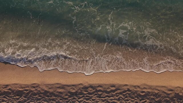 Top down drone view of sea waves gently breaking onto a sandy beach. White foam patterns and rhythmic surf create a peaceful coastal scene by the sea.