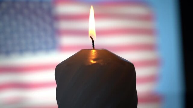 Candle flame flickers softly against blurred flag backdrop. Warm glow casts gentle shadow on dark wax surface. Symbolic stillness evokes remembrance or solemn reflection. Ideal for patriotic, memorial