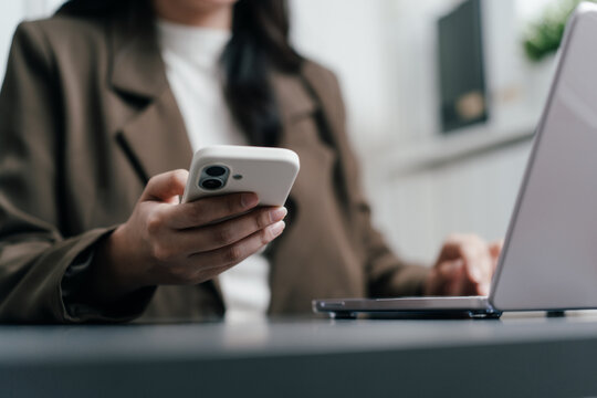 Businesswoman holding smartphone while working on a laptop. Professional female worker using mobile phone and digital technology for online business, AI technology and freelance work