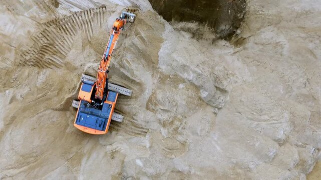 Aerial view shows an excavator digging into a sandy hillside. Orange machinery moves soil with its long arm and bucket. Deep tracks mark the dirt around the construction site