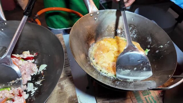 Thai Street Food Vendor Simultaneously Cooking Omelet and Stir-Fried Pork in Traditional Woks
