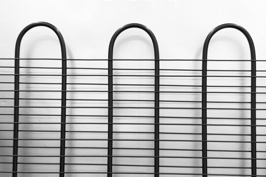 Black metal refrigerator condenser coils forming a horizontal and vertical grid pattern on a white wall