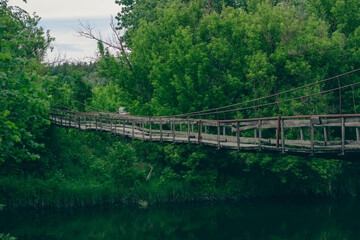 Fototapeta premium An old wooden suspension bridge stretching across a calm river, surrounded by lush green forest trees and dense vegetation under a cloudy sky. Concept of journey, pathway, adventure