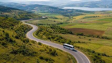 Fototapeta premium A white coach bus drives along a winding road through green hills, farmland and a lakeside valley, captured in an aerial panoramic landscape view
