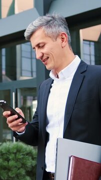 Man going outdoor with mobile phone in his hands. Middle aged businessman walking using smartphone outdoors in city near office building. Slow motion.