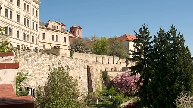 A ground-level view of the historic stone fortification walls and terraces in Litoměřice on a bright spring day. The scene features historic buildings and the red towers of a baroque church rising beh