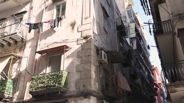 A low-angle view looking into a narrow, sunlit street characteristic of an Italian city, such as Naples. Tall, weathered residential buildings with balconies feature laundry hanging on clotheslines