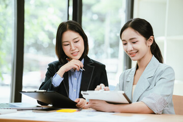 Obraz premium Two businesswomen collaborating at a modern office desk, discussing a project while reviewing information on a laptop and tablet in a bright professional workspace.