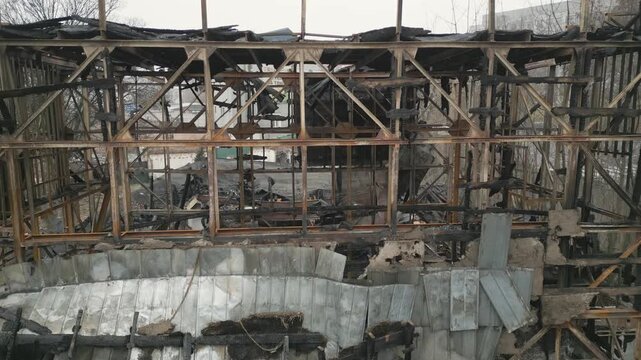 An aerial close-up view reveals the charred metal and wooden skeletal frame of a building devastated by fire. The structure shows severe damage with burnt beams, ash, and collapsed roofing components.