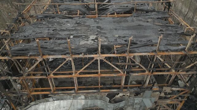 An aerial close-up view reveals the charred metal and wooden skeletal frame of a building devastated by fire. The structure shows severe damage with burnt beams, ash, and collapsed roofing components.