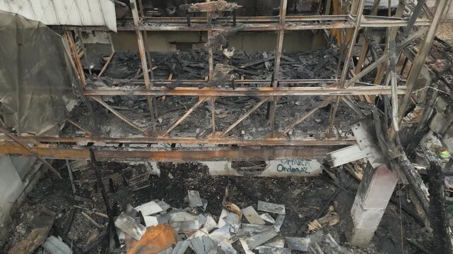 An aerial close-up view reveals the charred metal and wooden skeletal frame of a building devastated by fire. The structure shows severe damage with burnt beams, ash, and collapsed roofing components.