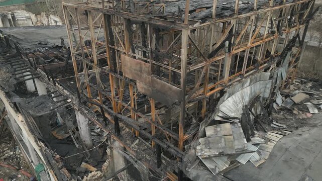 An aerial close-up view reveals the charred metal and wooden skeletal frame of a building devastated by fire. The structure shows severe damage with burnt beams, ash, and collapsed roofing components.