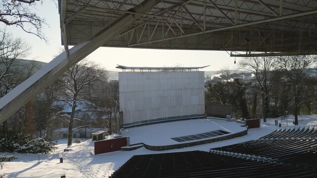 An elevated drone winter view of the open-air summer cinema in &Uacute;st&iacute; nad Labem shows rows of empty, snow-covered amphitheater seating and a large projection screen under a massive metal roof structure 