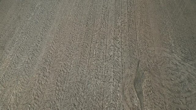 Aerial drone view captures a vast, empty agricultural field with freshly plowed dry soil. The surface shows textured tractor tire tracks and linear cultivation lines stretching across the bro
