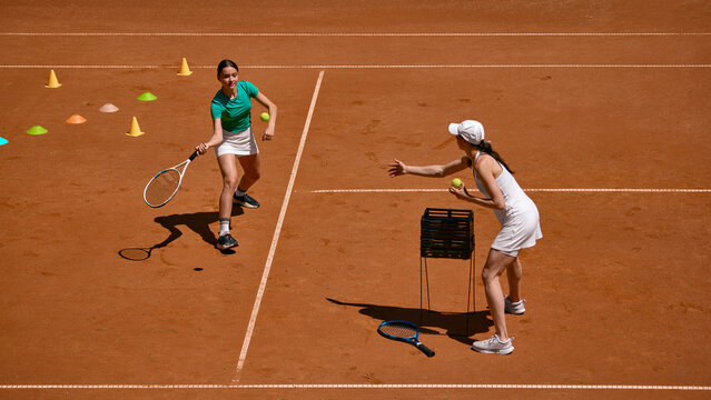 Tennis coach feeds ball to teenage girl during forehand drill. Concept of private lesson, court workout, beginner progress, sports discipline and teen fitness.