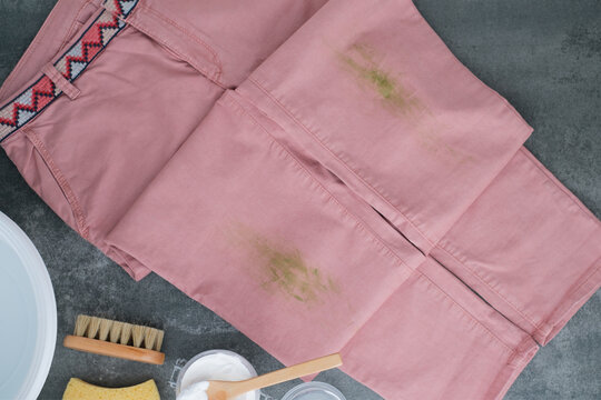 Pink jeans with green stains rest on a gray background. Cleaning tools including a wooden brush, baking soda, sponge, and soap are nearby. Concept eco-friendly cleaning products