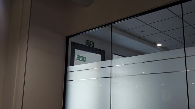 Zoom-in footage of fire exit sign behind glass office partitions, ideal for workplace safety training, emergency evacuation planning, corporate compliance, and office management projects.