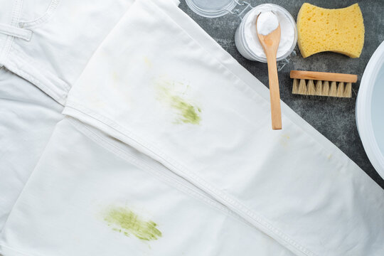 White jeans with green stains rest on a gray background. Cleaning tools including a wooden brush, baking soda, sponge, and soap are nearby. Concept eco-friendly cleaning products