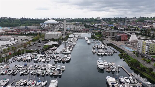 Hundreds of boats fill Thea Foss Waterway marina with Tacoma Dome and cable-stay bridge rising beyond colorful waterfront apartment buildings in Tacoma, Washington, USA. Aerial drone flyover shot,