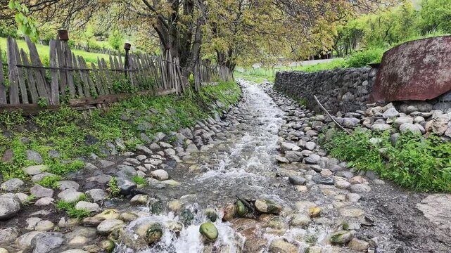Small mountain stream flowing over rocky bed between wooden fence and stone wall in lush green countryside setting, Mestia, Svaneti, Georgia