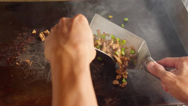 Close up video of a cook using a metal spatula to chop and stir seasoned beef with green paprika on a hot flat top griddle