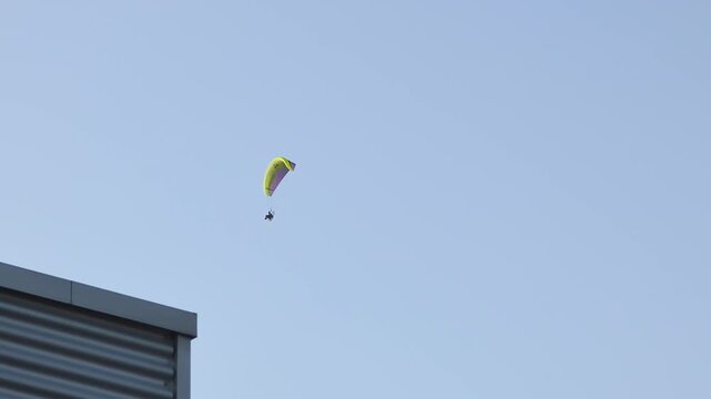 Paramotor powered paraglider flying above buildings during sunny day in clear sky