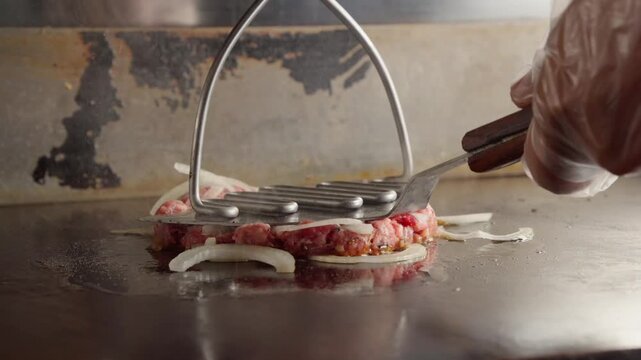 A chef pressing a ball of ground beef topped with thinly sliced raw onions onto a hot griddle to create a smash burger