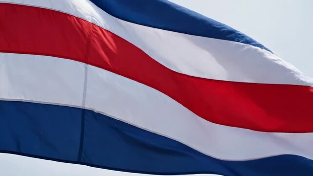 Costa Rica flag, horizontal blue, white red stripes waving in slow tracking medium close-up, pale clear sky in bright daylight. costa rican national pride and central american identity