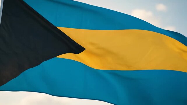 Bahamas national flag waving against cloudy sky, bold black triangle on left with aquamarine blue stripes and golden yellow horizontal band, silky fabric in motion. Concept of bahamian national pride 