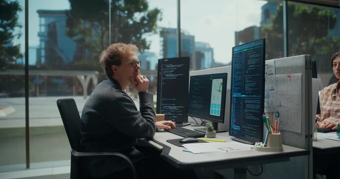 Young Caucasian Male Software Developer Codes a New Mobile App on Multiple Monitors in a Tech Office, Focused on Programming Beside a Female Colleague at a Busy Startup. Working Using Computer