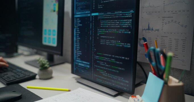 Portrait of Male Software Developer Types Code on a Keyboard Beside Multiple Monitors Showing Programming Scripts, Mobile App Interfaces, Analytics, Driven Startup Office. Working Using Computer
