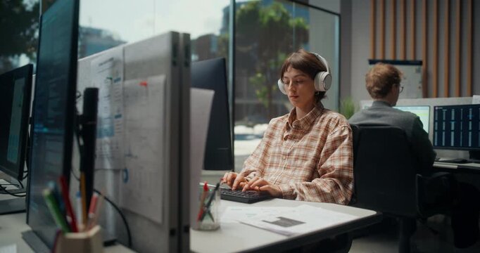 Portrait of Female App Engineer in Types at a Workstation, Reviews Source Code on Monitor. Teammates Analyzes Dashboards in Modern Software Development Studio. Working Using Computer